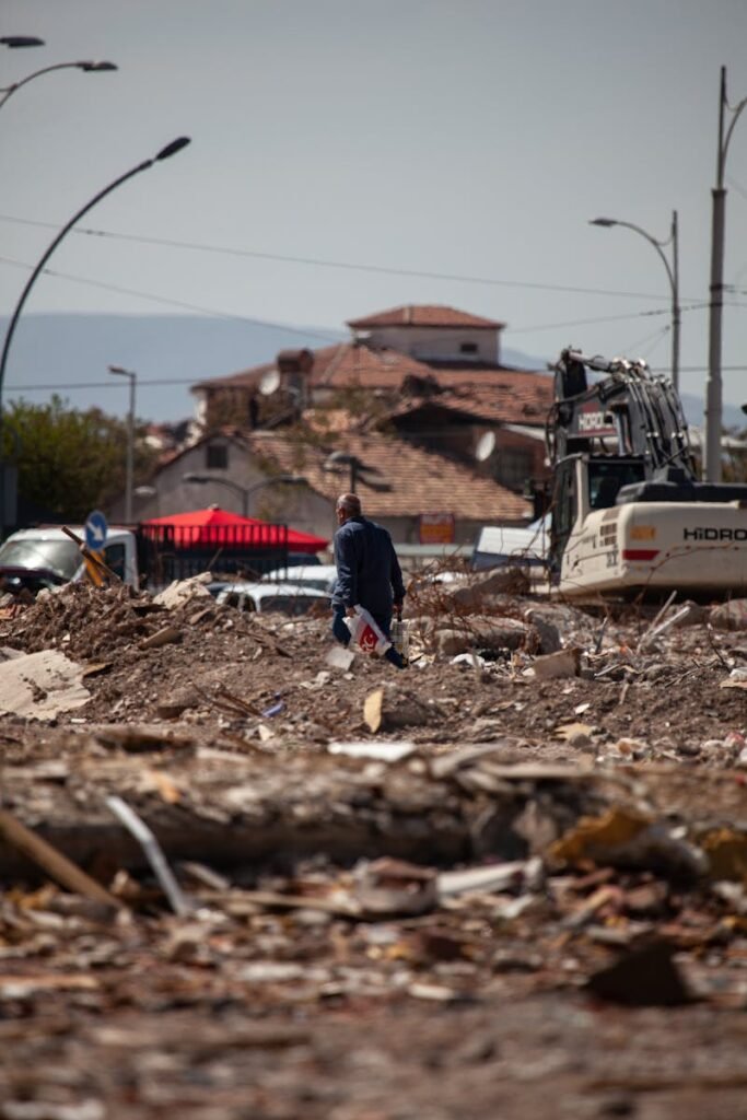 A man walks through a demolished urban area in Malatya, Türkiye, amidst debris and construction equipment.